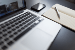 Black desk with laptop, open notebook, and smart phone on top of it.