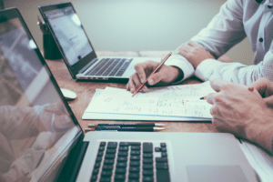 Two people working at a desk, looking at a piece of paper with two laptops.