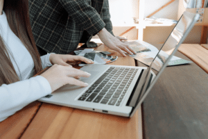 A man and a woman working on a laptop computer and a wooden desk.