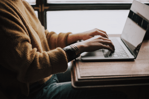 Person in a yellow sweater sitting at a desk with a laptop righting a customer review. 